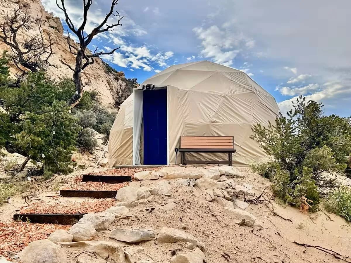 The geodesic dome at Luxe Den Capitol Reef, white shell with a blue door, on a stone path below the sandstone cliff