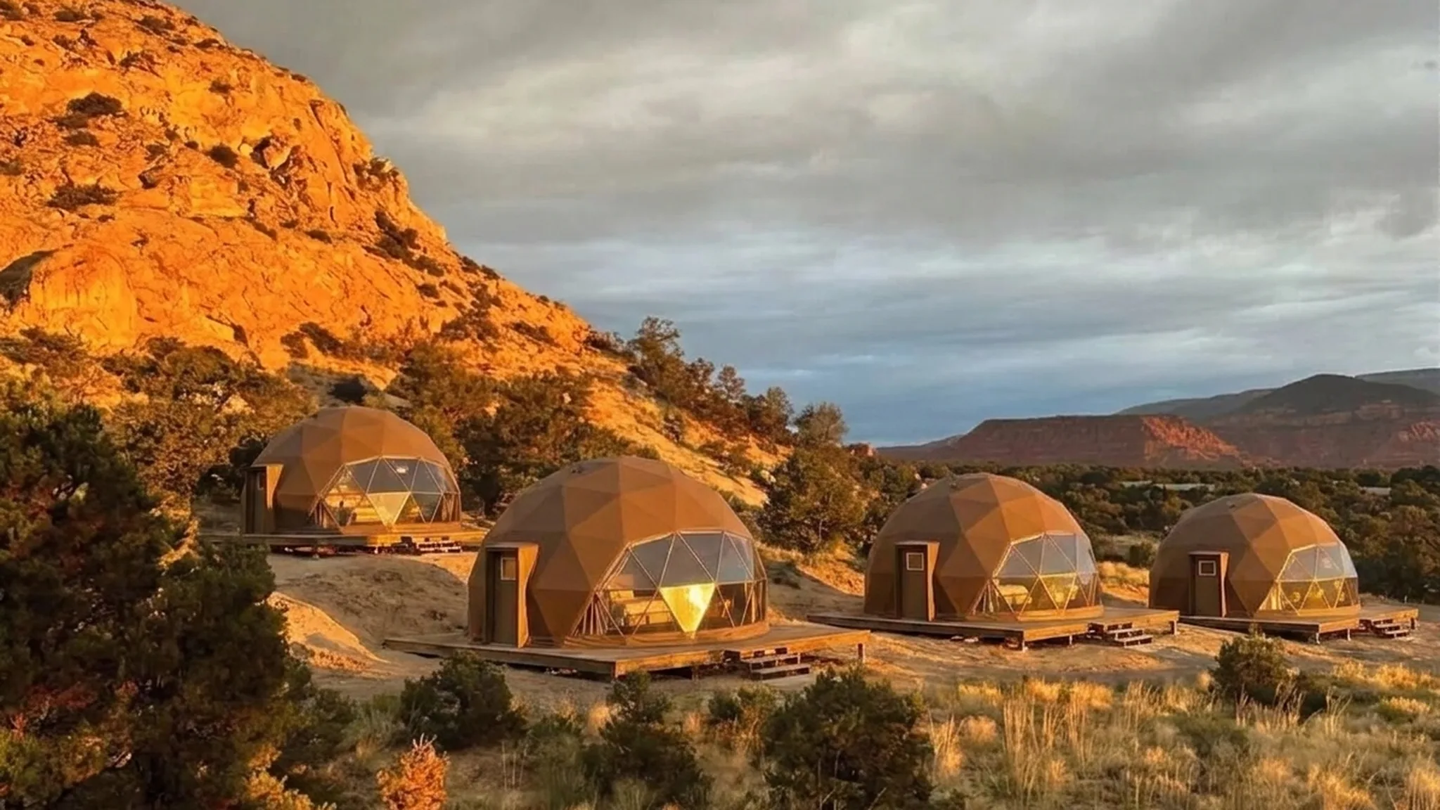 Visualisation of four brown geodesic domes on a sand bench below a sandstone slope near Capitol Reef, low golden light warming their panels