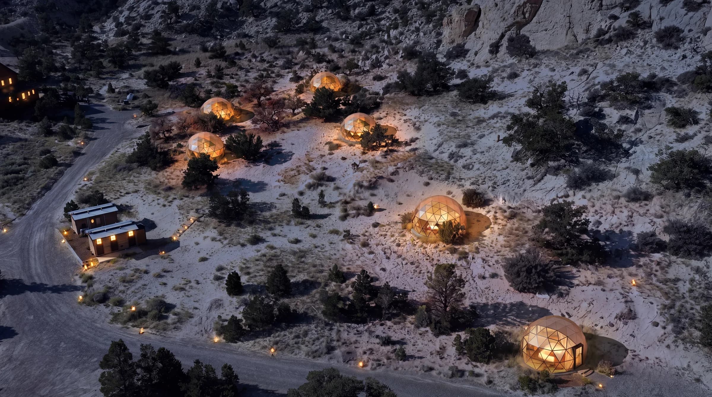 The same six planned glass domes at night, lit warm from inside, with path lights along the gravel roads