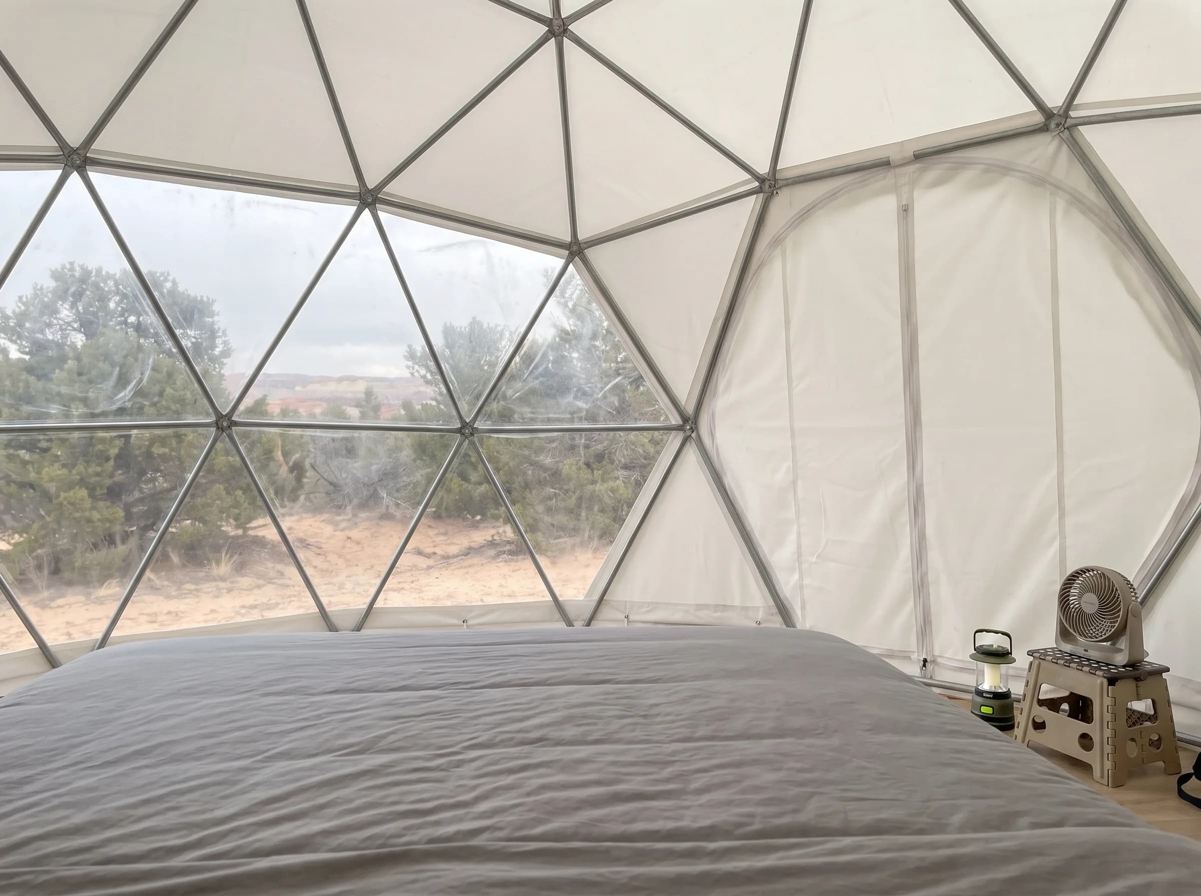 Visualisation of a typical PVC-fabric glamping dome interior: a grey bed facing a cloudy, scuffed vinyl window with the desert soft and hazy behind it