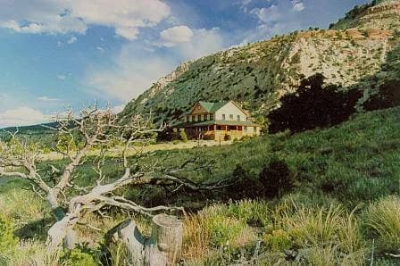 The Muley Twist Inn from the meadow below, a two-storey timber building under the sandstone ridge, with a dead juniper in the foreground