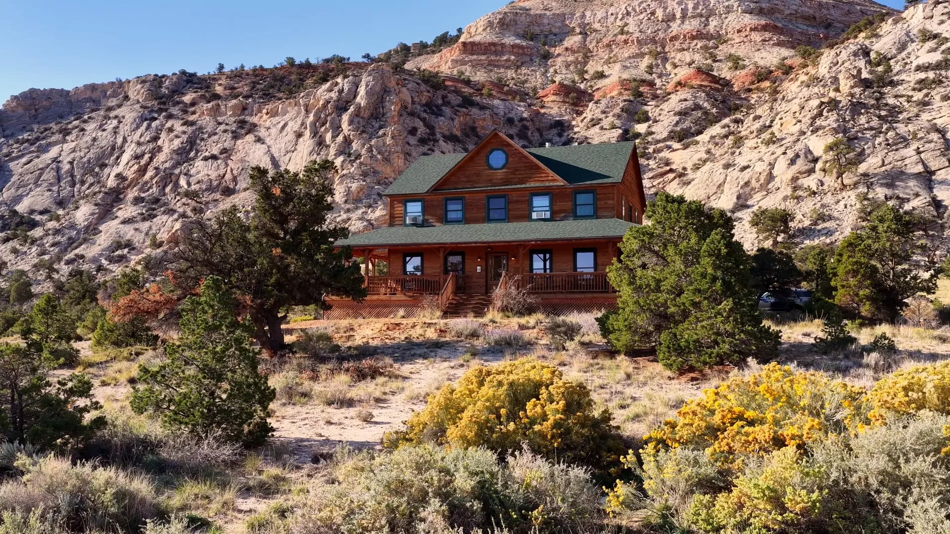 Aerial view of The Lodge, a two-storey timber building with a green roof and a wraparound deck, among junipers and rabbitbrush below white and red sandstone cliffs near Capitol Reef National Park