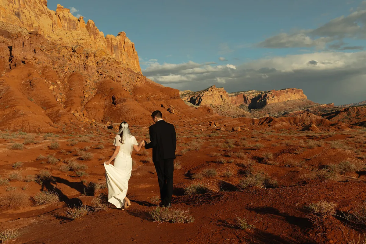 A bride and groom walking across red rock below Capitol Reef cliffs